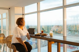 Una persona sentada a una mesa con una computadora portátil, un sándwich y una bebida en una habitación con ventanas amplias.
