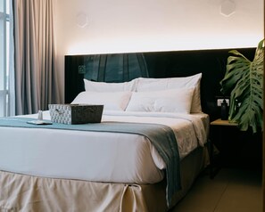 Habitación de hotel con cama, mesita de noche, planta y ventana con cortinas.