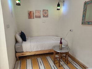 Een knus kamertje met een enkel bed, een klein tafeltje en een wandlamp.
