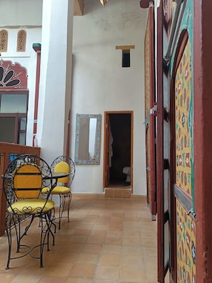 A corridor with a tiled floor, a wooden door, a mirror, and a chair with a cushion.