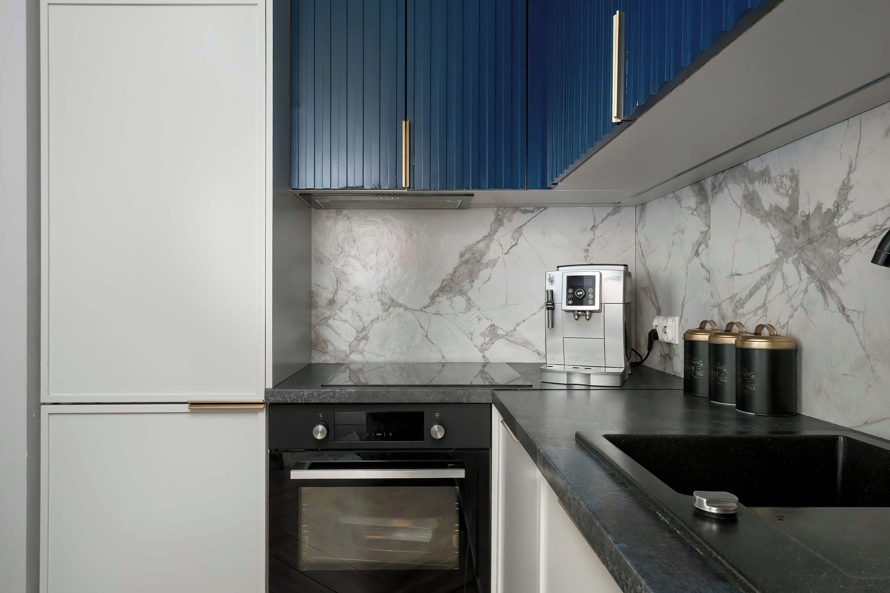 A sleek kitchen is outfitted with deep blue upper cabinets, a marble backsplash, and white lower drawers. A built-in oven and an espresso machine are perfectly integrated into the black countertop workspace.