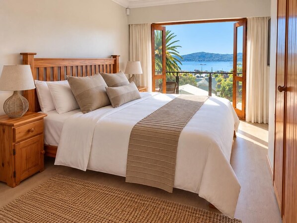 A bedroom with a large bed, wooden headboard, two bedside tables with lamps, a woven rug, and a view of the sea through a glass door. - 6 Bond St (Knysna)