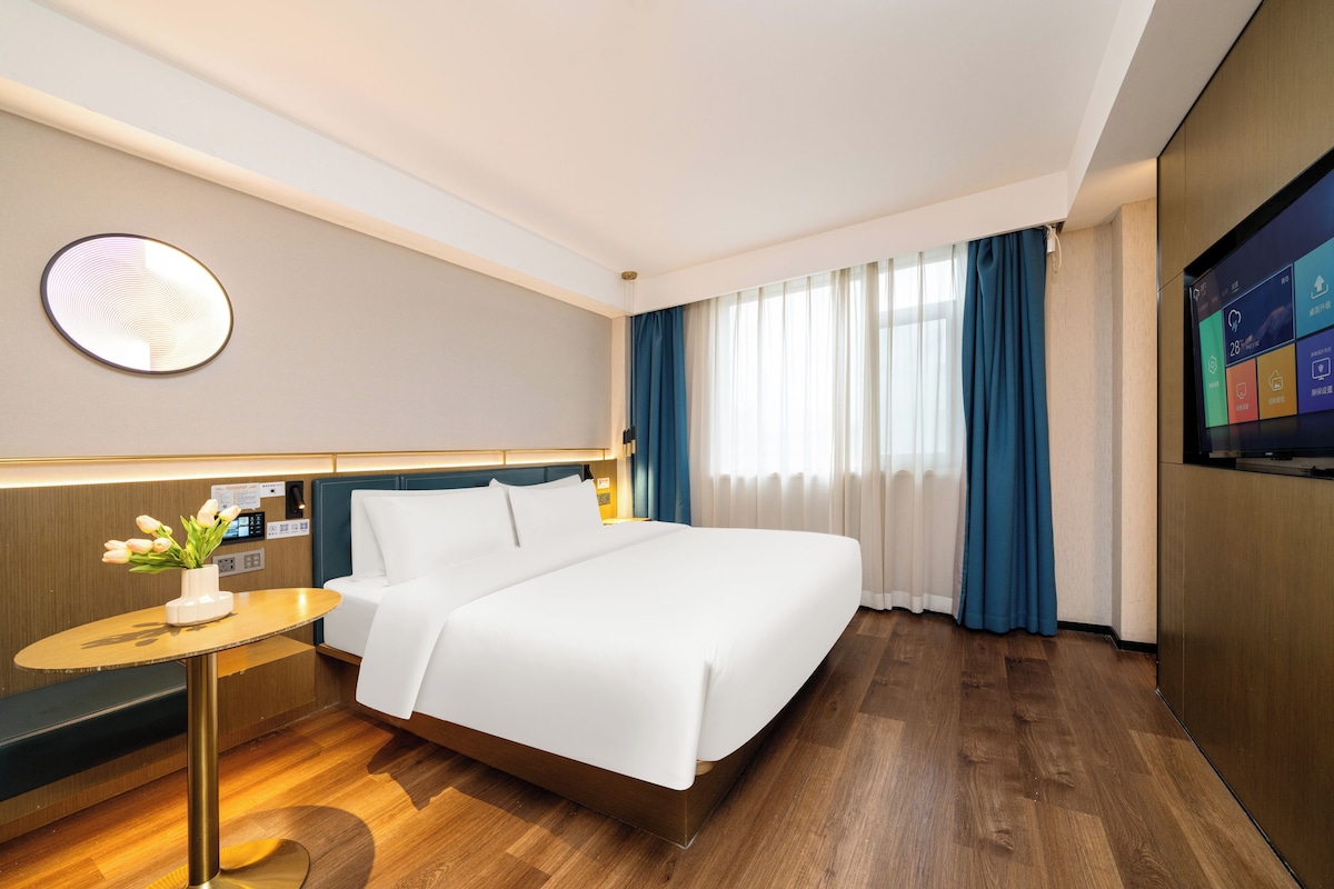 Una habitación de hotel con cama, escritorio con lámpara, reloj de pared, televisor y ventana con cortinas.
