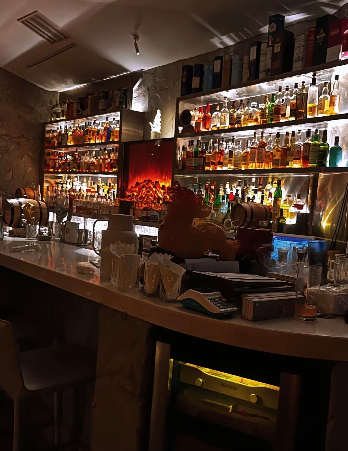 Zona de bar con estantes llenos de botellas de licor, una caja registradora y un monitor de ordenador.