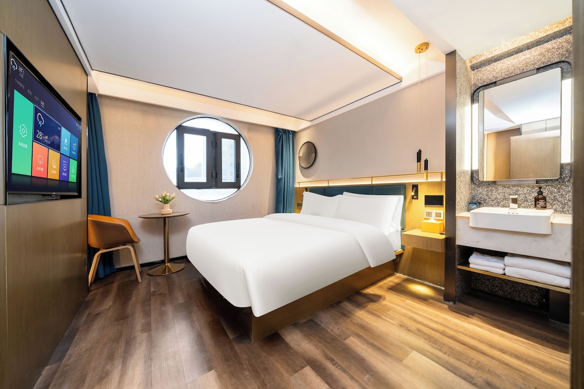 Habitación de hotel con cama, ventanal redondo, una mesita con un florero, espejo circular, mesita de noche con lámpara y baño con lavamanos y toallero.