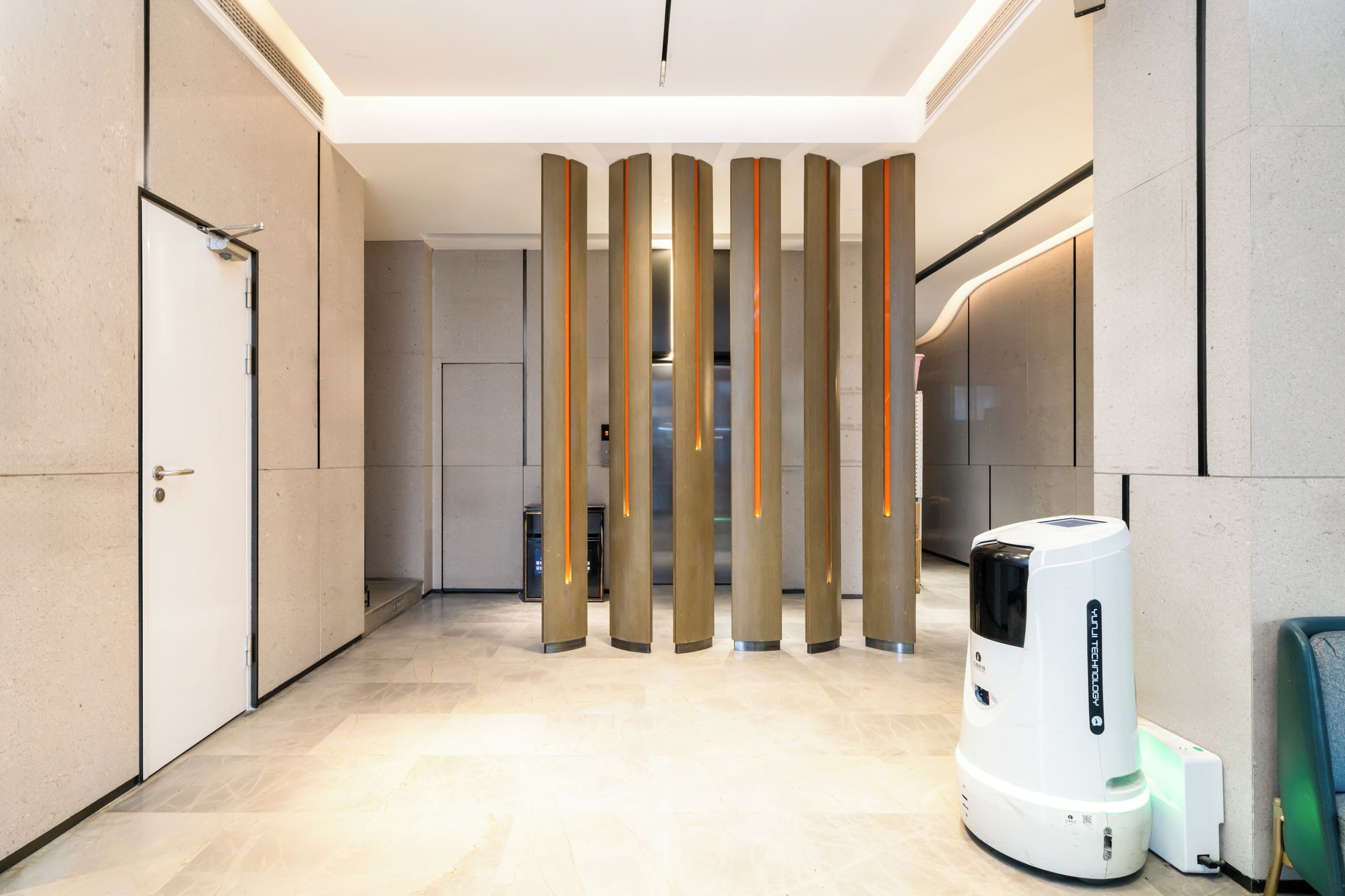 Un pasillo moderno con una serie de paneles verticales, un aspirador robot blanco y una puerta a la izquierda.
