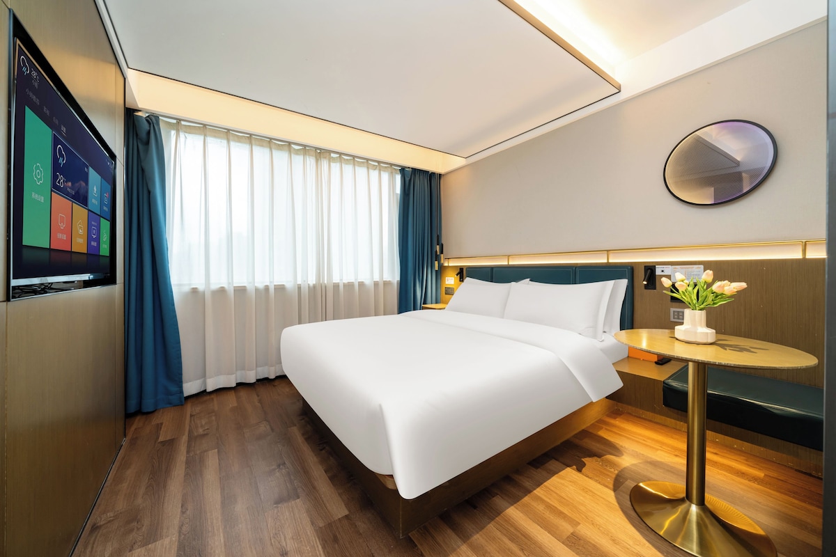 Habitación de hotel con cama, mesita de noche con lámpara, espejo redondo y televisor empotrado mostrando la temperatura ambiente.