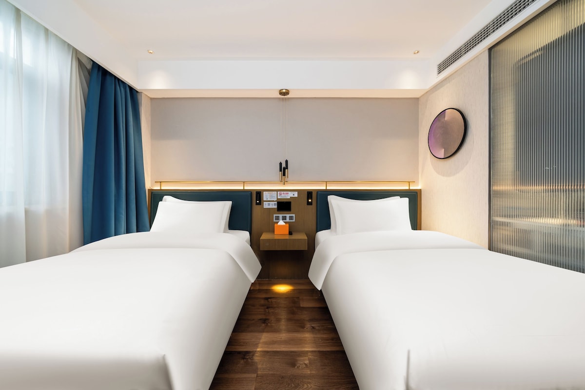 Habitación de hotel con dos camas, piso de madera, una mesita de noche con lámpara y vista a la ciudad por la ventana.