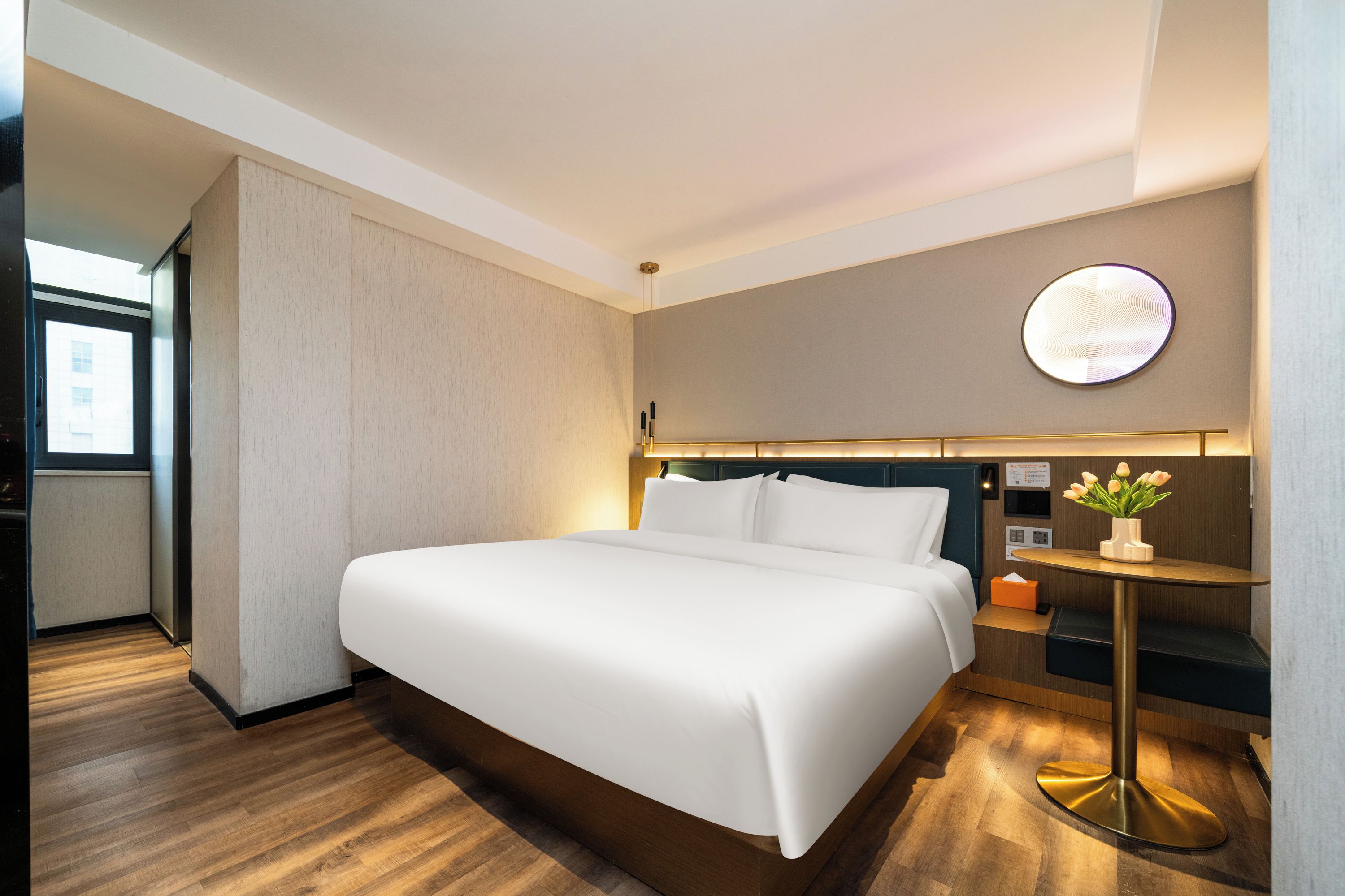 Habitación de hotel con una cama grande, una mesita de noche con un florero, una lámpara de pared y vistas al exterior.