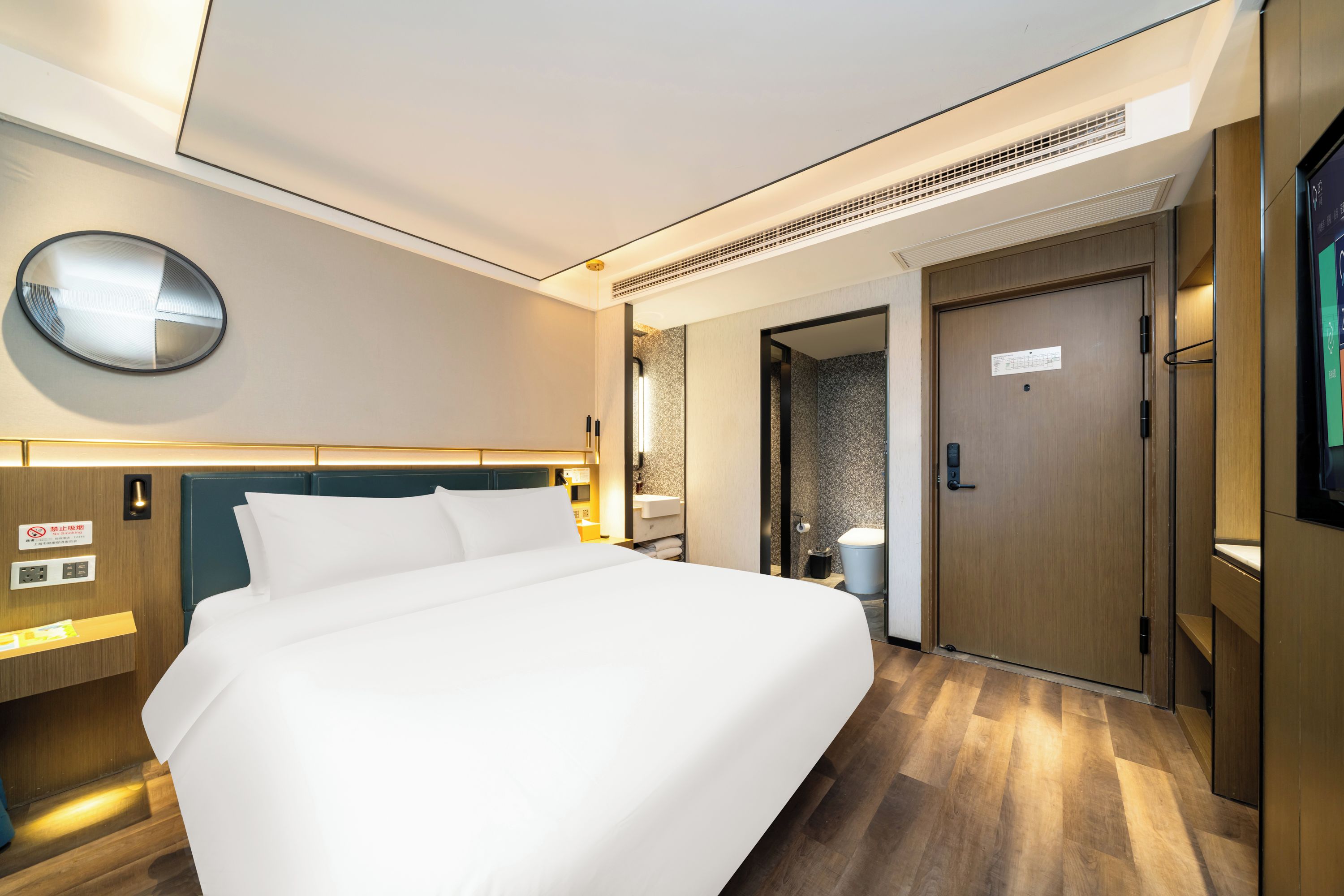 Een hotelkamer met een groot bed, een ronde spiegel, een houten deur en een aan de muur bevestigde televisie.