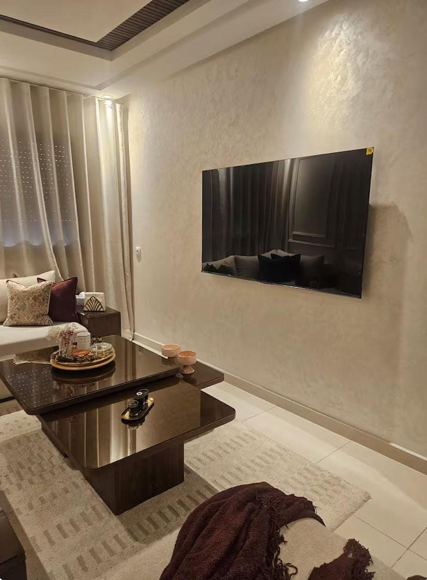 Un salon moderne avec un téléviseur à écran plat fixé au mur, une table basse de couleur sombre et un canapé agrémenté de coussins.