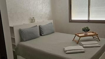 A bedroom with a bed, pillows, a bedside table with a plant, a window with blinds, and a decorative ceiling light.