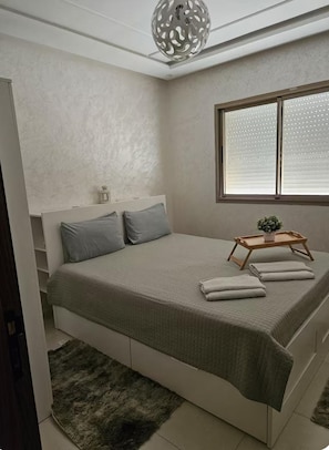 A bedroom with a bed, pillows, a bedside table with a plant, a window with blinds, and a decorative ceiling light. - New Listing (Kénitra)
