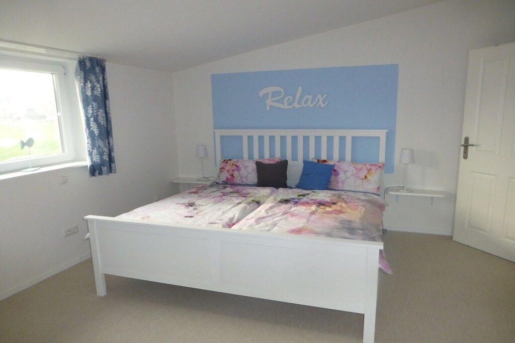 Une chambre à coucher avec un lit, une fenêtre avec des rideaux, un mur orné d’une inscription « Détendez-vous » et une porte.