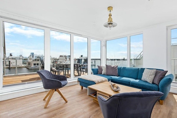 A modern living room with a sofa, chairs, and a coffee table. Large windows provide a view of the cityscape and water.