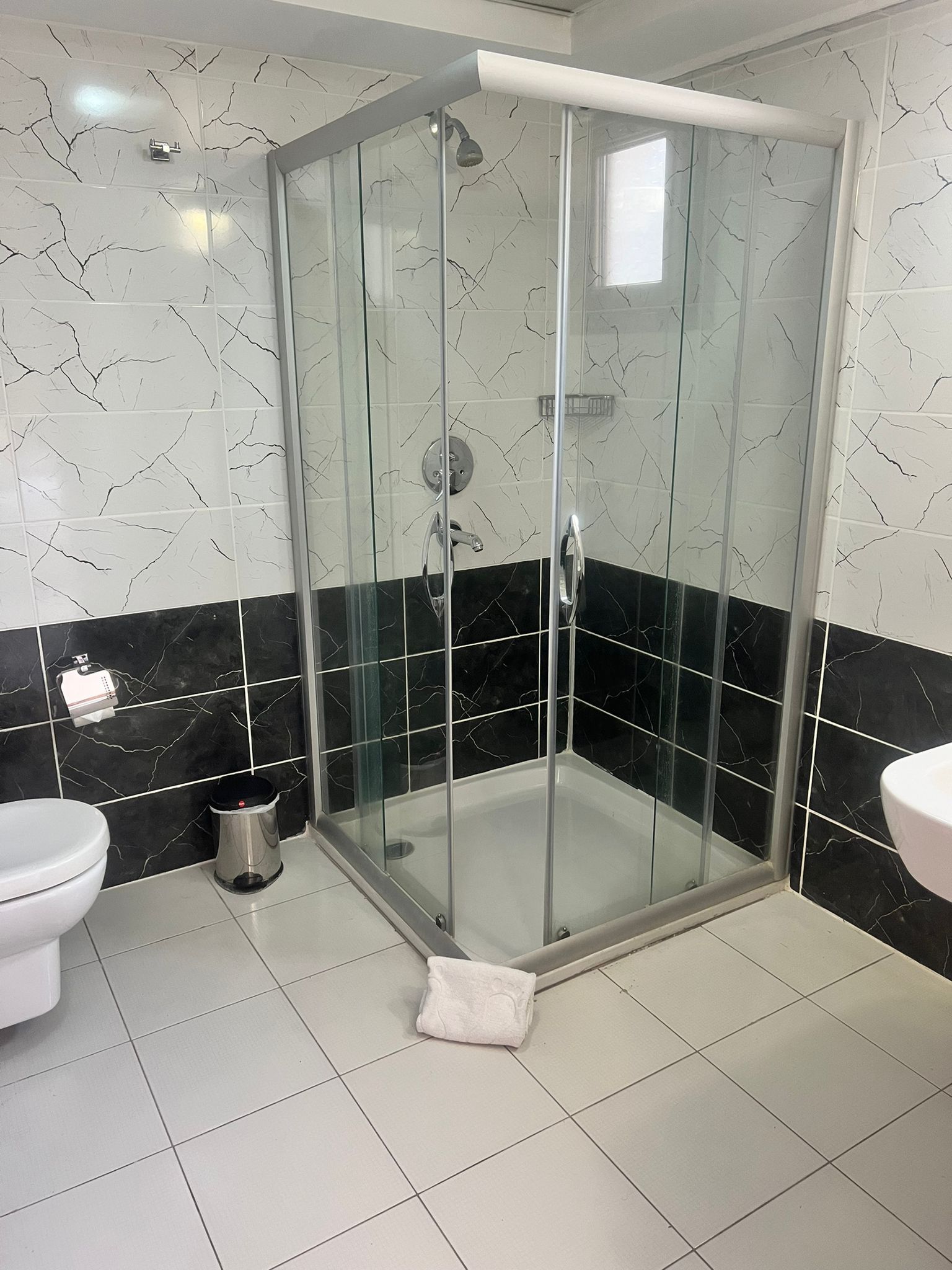 Un bany amb dutxa tancada amb vidre, parets revestides de rajoles blanques i negres, un vàter blanc i un lavabo blanc.