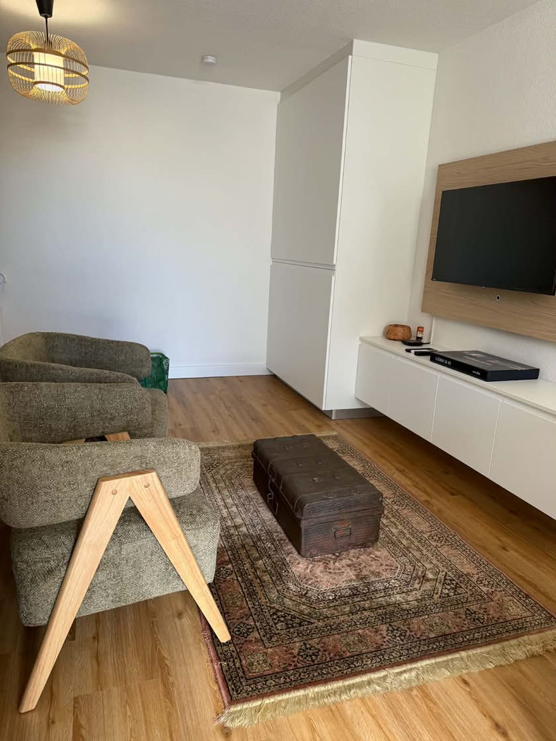 A modern living room with a sofa, a wooden stool, a patterned rug, a television mounted on the wall, and a wooden floor.