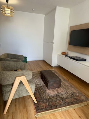 A modern living room with a sofa, a wooden stool, a patterned rug, a television mounted on the wall, and a wooden floor. - New Listing (Saint-Denis)