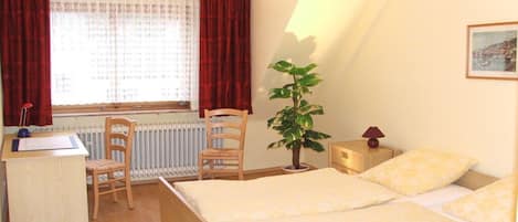 A bedroom with a bed, a desk, a chair, a radiator, a potted plant, and a lamp.