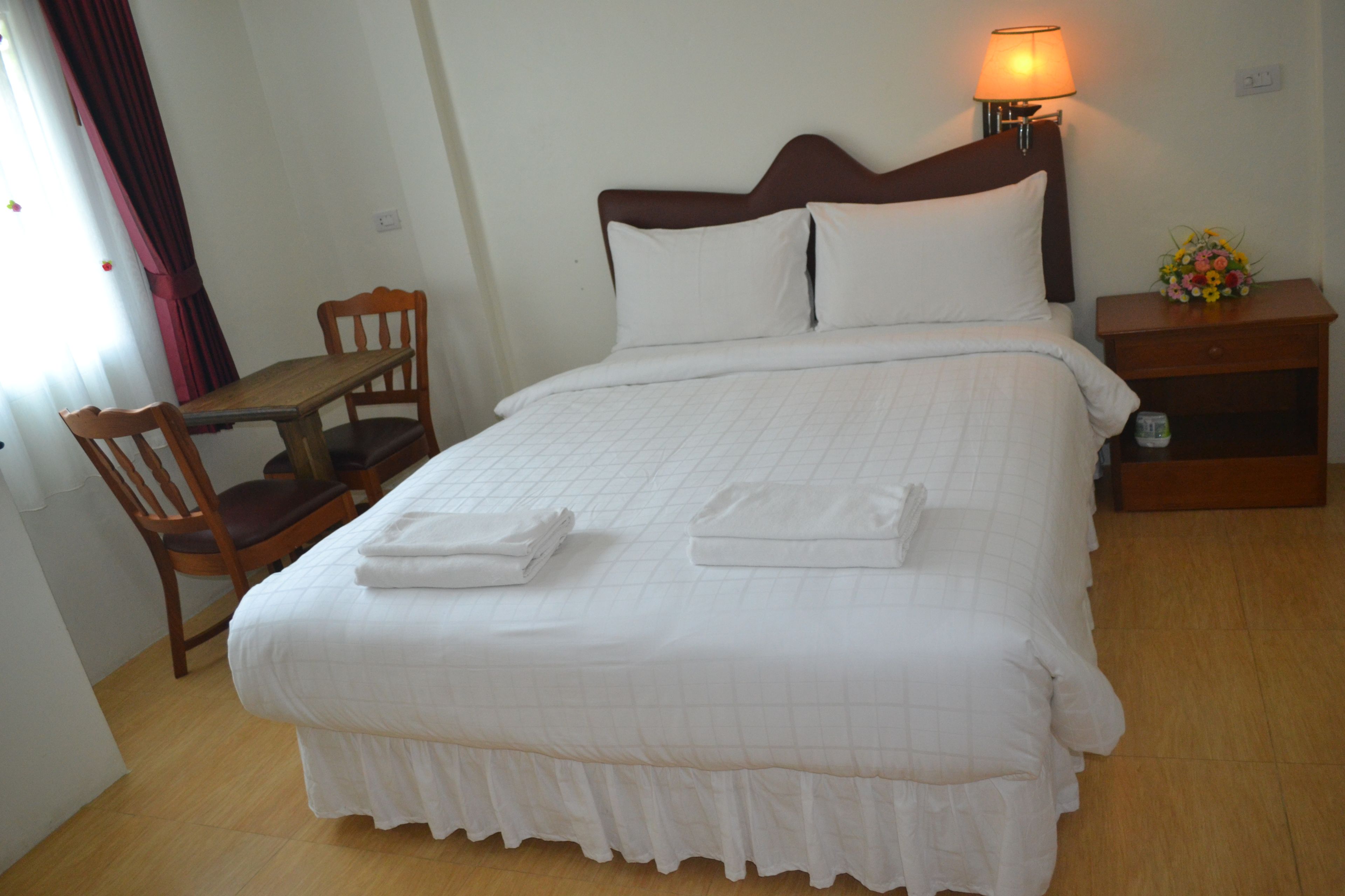 Una cama bien hecha con sábanas blancas, una mesita de noche con una lámpara, una silla de madera y una pequeña mesa con un florero.