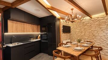 Une cuisine moderne avec des meubles de couleur noire, une table à manger en bois pour quatre personnes et un mur en pierre agrémenté d’une cheminée.