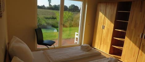 A bedroom with a bed, a chair, a wooden wardrobe, and a sliding glass door to a garden.