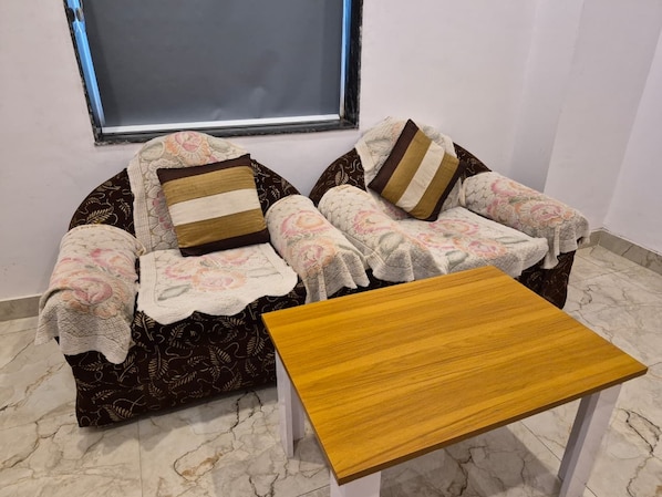 A two-seater sofa with a floral cover and patterned cushions, a wooden coffee table, and a window with a view.