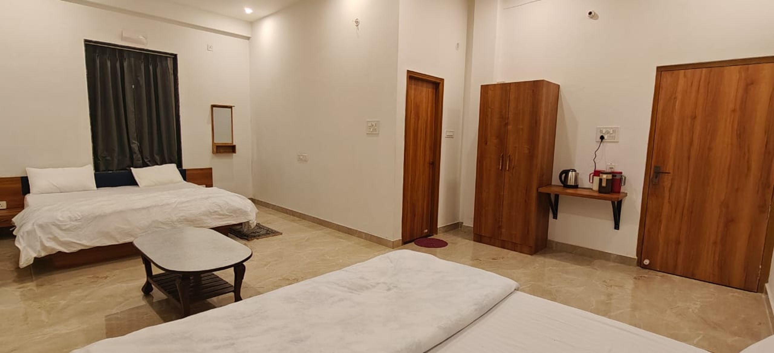 Une chambre d’hôtel avec deux lits, une petite table, un miroir, une armoire et une étagère avec une bouilloire.