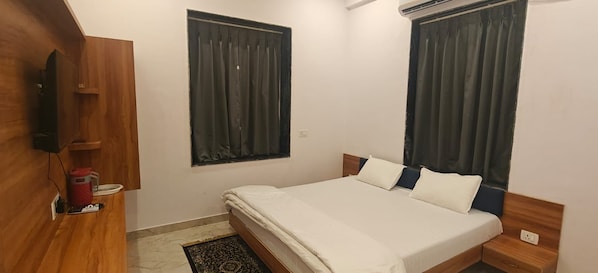A bedroom with a bed, a wooden wardrobe, a TV, and two windows with curtains.