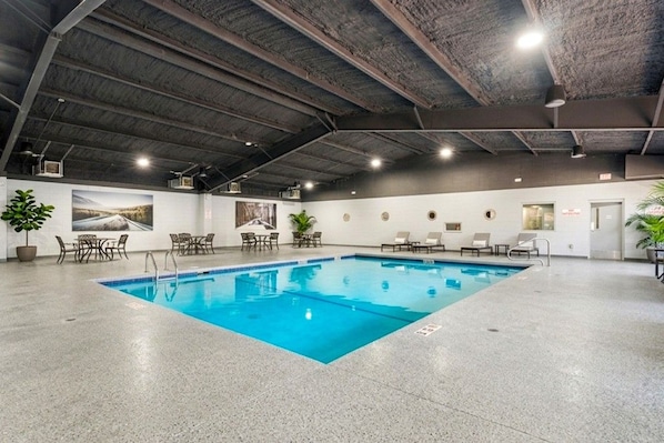 Una piscina interna con acqua cristallina, circondata da lettini e tavolini, sotto un soffitto alto con travi a vista.