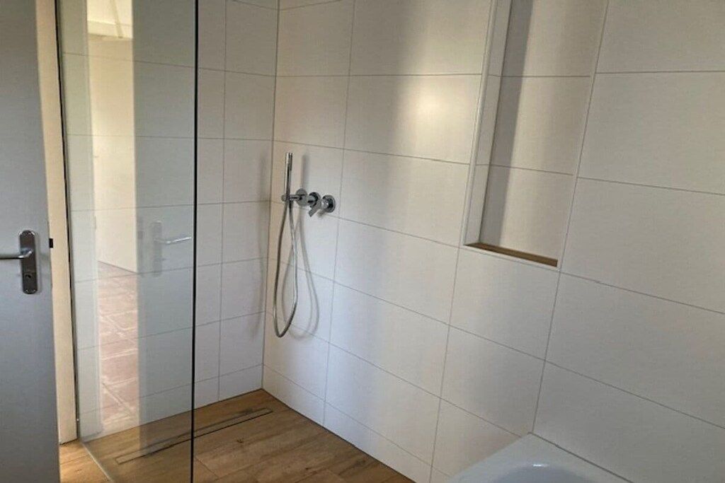 A bathroom with white tiled walls, a glass shower enclosure, and a wooden floor.