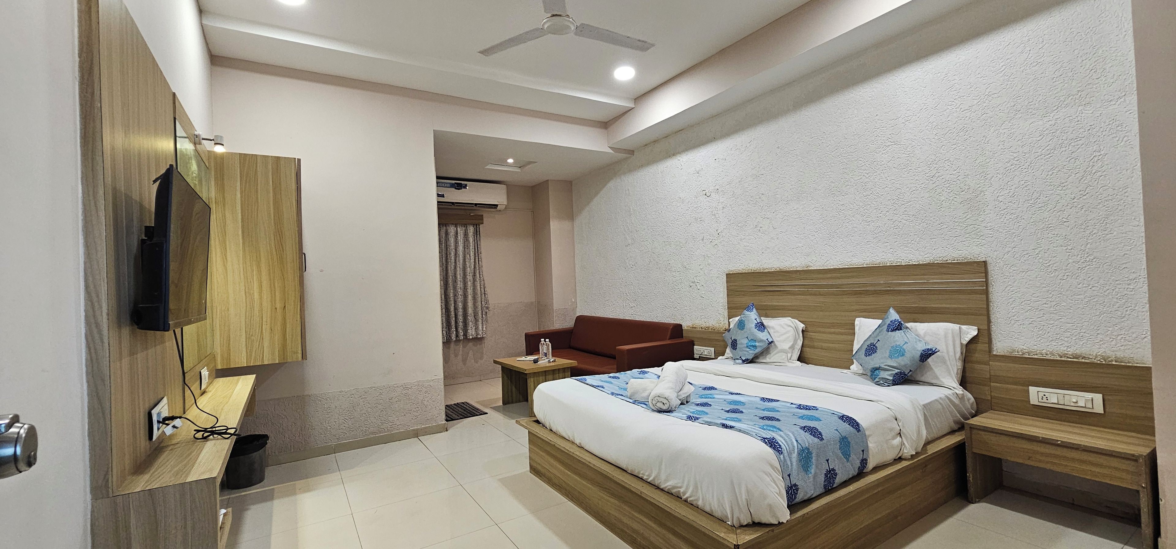 Una habitación de hotel con cama, cabecera de madera, mesita de noche, televisor montado en la pared, una pequeña mesa y una silla.