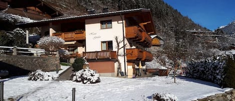 A chalet-style building with a wooden facade, a balcony, and a stone wall in a snowy mountainous area.