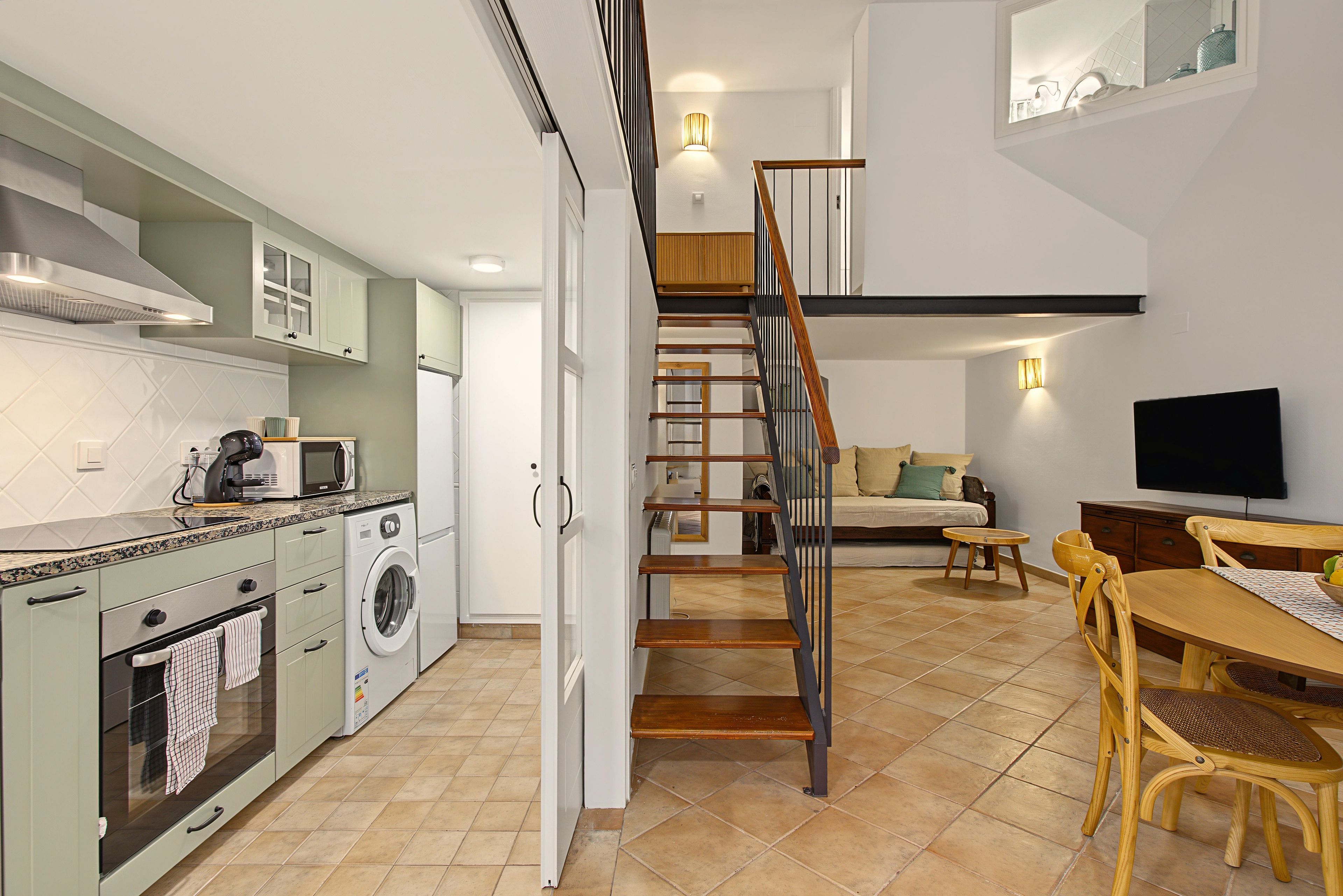 A modern kitchen with a built-in washing machine, a dining area with a wooden table and chairs, and a staircase leading to an upper level.