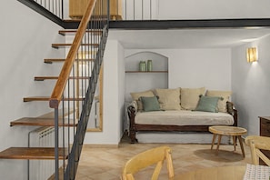 Ein Zimmer mit einer Holztreppe, einer Couch mit Kissen, einem kleinen runden Tisch und einem Sessel.