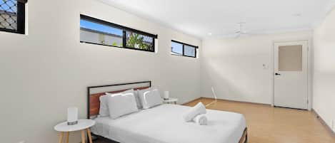 A modern bedroom with a bed, bedside tables, a ceiling fan, and windows with views.
