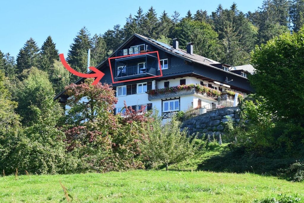 Una casa de varios pisos con balcón, rodeada de árboles y un muro de piedra.