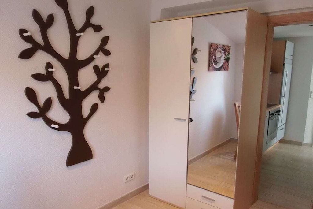 Une pièce avec un placard, un décor mural représentant un arbre et un coin cuisine visible à travers une porte ouverte.