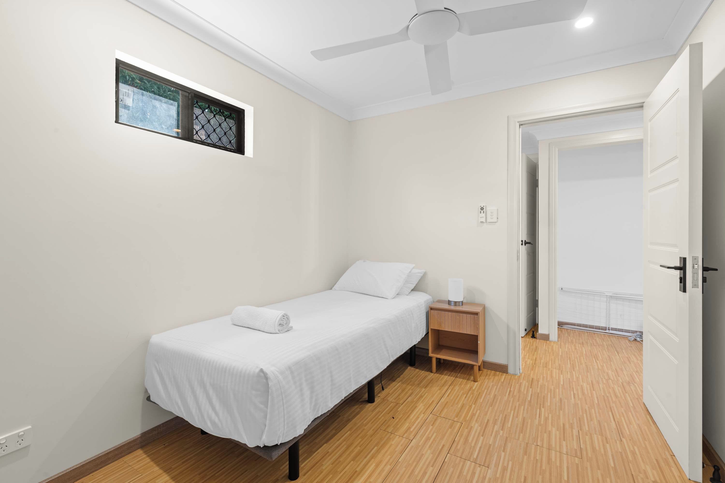 A single-bed room with a ceiling fan, a small bedside table with a lamp, and a door leading to another room.
