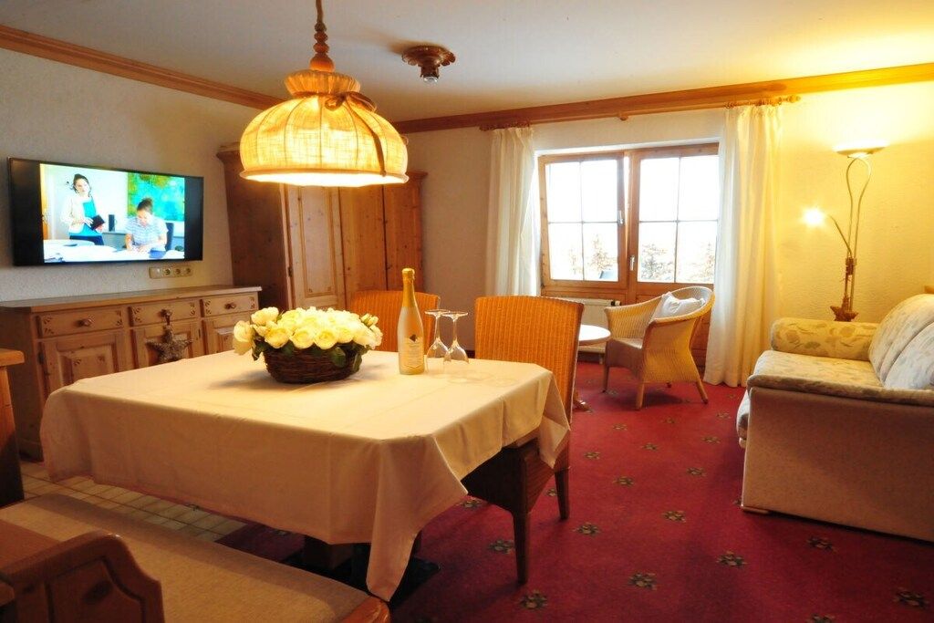 A dining area with a table set for two, a television mounted on the wall, and a window with curtains.