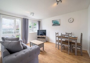 Ein Wohnzimmer mit grauem Sofa, einem Holztisch, einem Fernsehgerät auf einer Holzkommode, einem Esstisch mit Stühlen und einer Wanduhr.