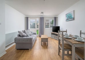 Uma sala de estar moderna com um sofá cinzento, uma mesa de centro de madeira e uma área de refeições com uma mesa e cadeiras de madeira.
