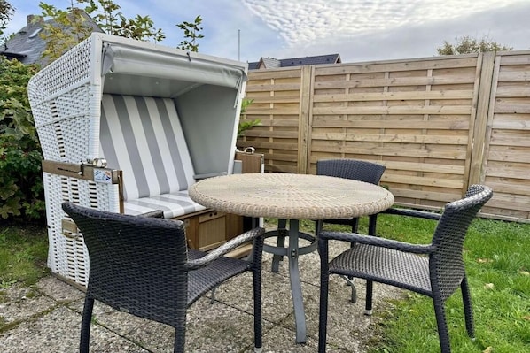 A wicker outdoor seating area with a round table and chairs, a cushioned bench, and a wooden fence in the background.