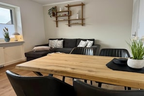 A modern living room with a wooden dining table, black leather chairs, a grey sofa with patterned cushions, and a wooden shelf with decorative items.