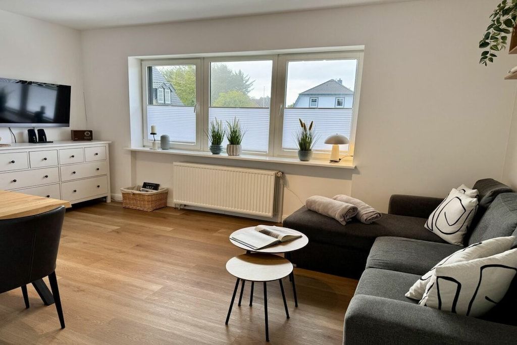 A modern living room with a sofa, a small table, a TV, and a window with plants.