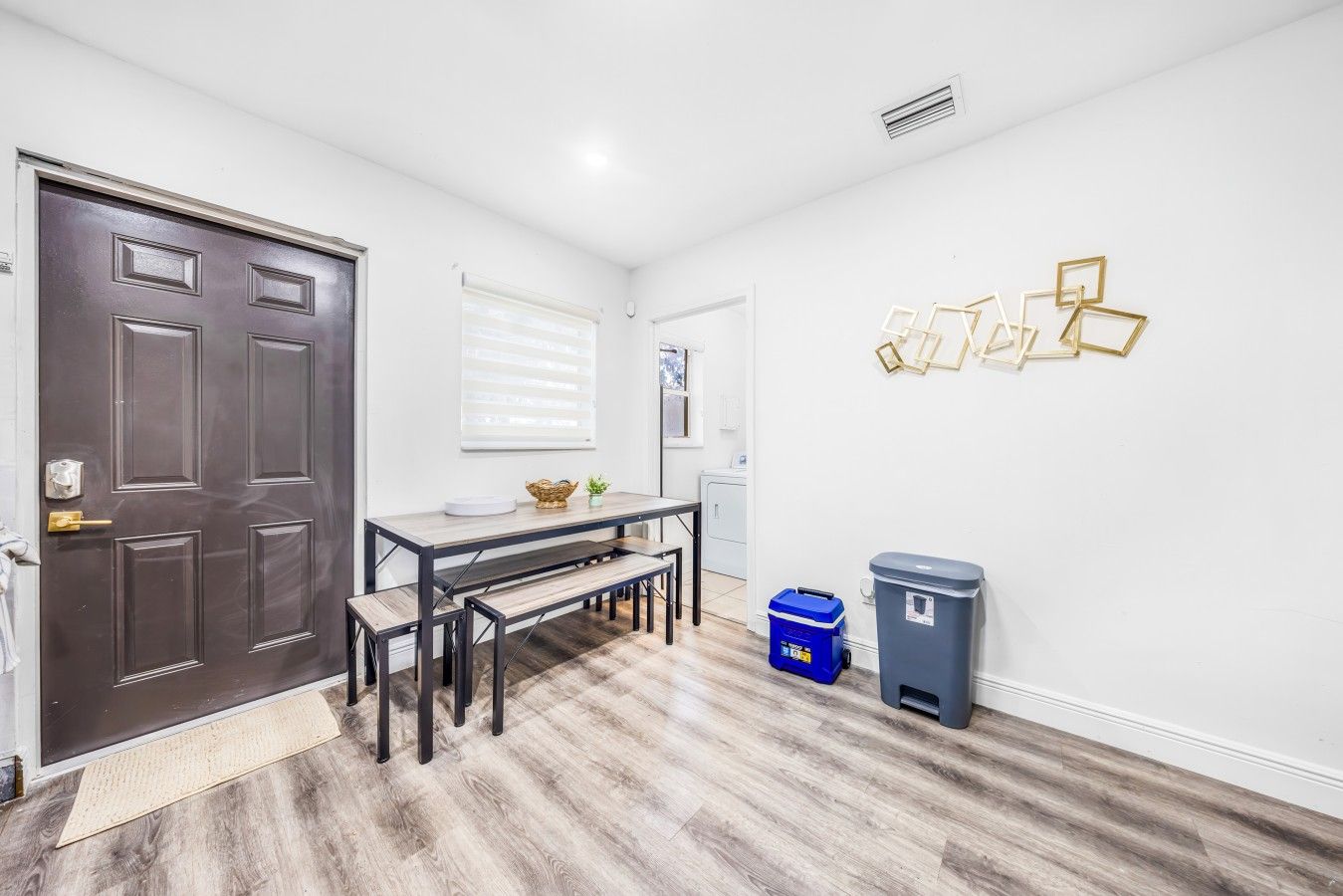 A room with a dark wooden door, a dining table with benches, a blue trash bin, and a gray wastebasket.