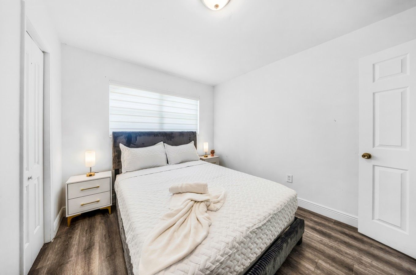 A bedroom with a bed, bedside tables, a window with blinds, and a door.