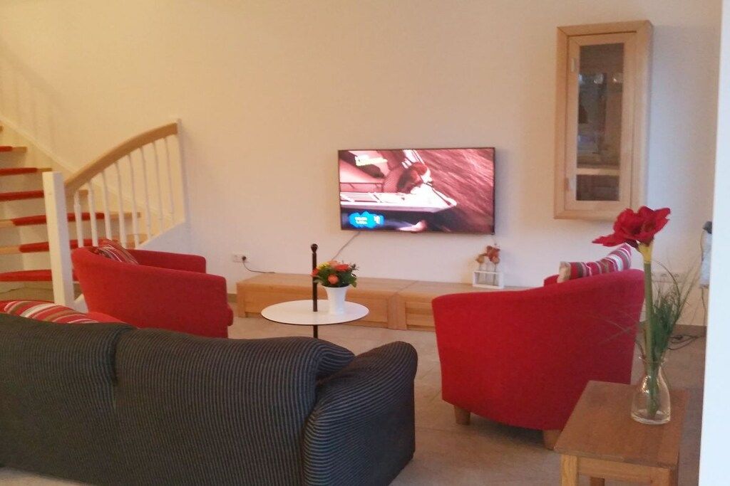 Un salon avec un téléviseur fixé au mur, un escalier et des fauteuils recouverts de tissu rouge.