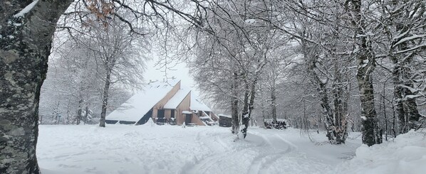 A snow-covered landscape with a building featuring a sloped roof, surrounded by trees.