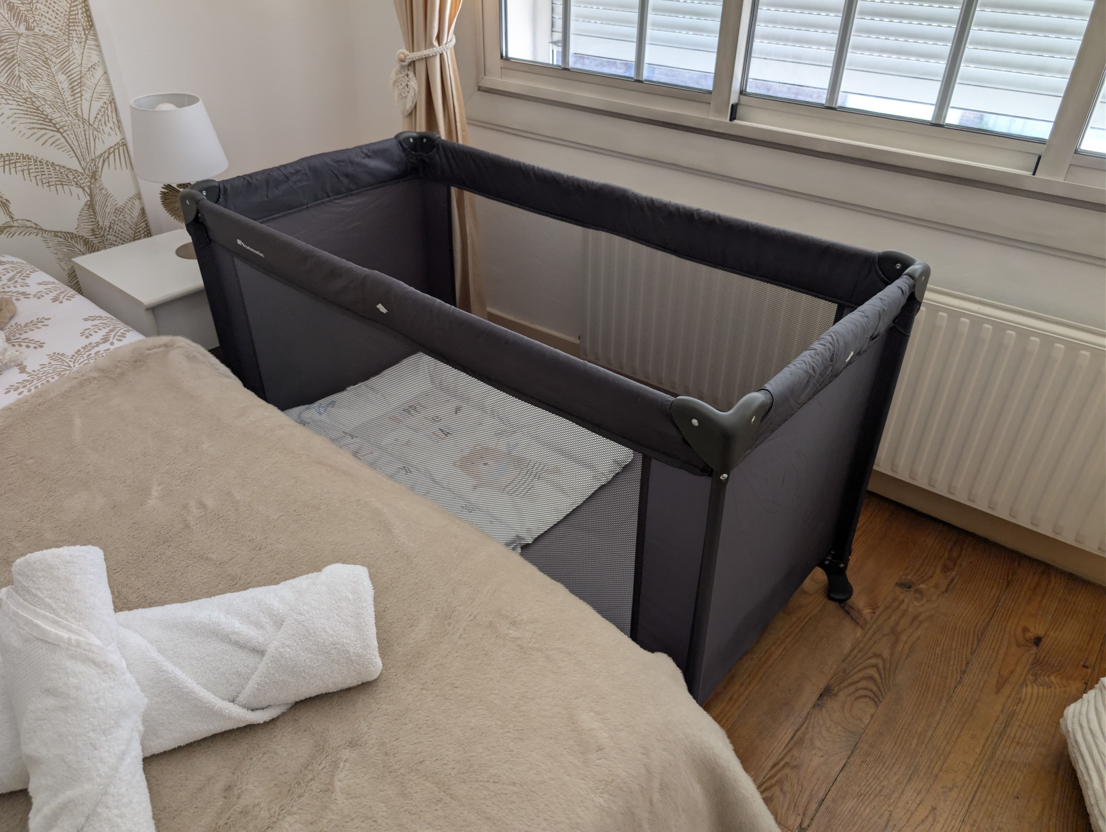 Une chambre avec un berceau, un lit recouvert d’une couverture beige, une serviette blanche, une fenêtre avec des stores et un radiateur.